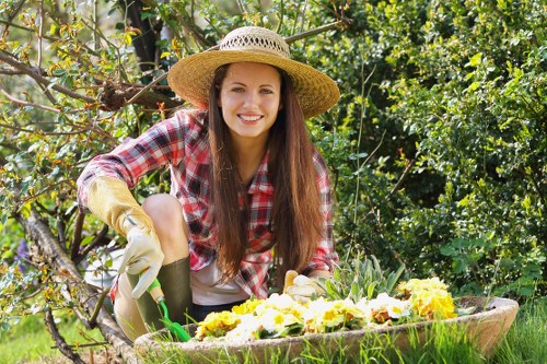 A young woman with long brown hair, wearing a straw sun hat, plaid shirt, gardening gloves, and beige trousers, is kneeling on a lush green lawn in a garden. She is smiling as she tends to a flower bed containing bright yellow and white flowers, with her left hand supporting the plants and her right hand holding a small gardening trowel. Behind her, there are shrubs and small trees with green foliage, and a wooden branch extends across the background, indicating a landscaped outdoor space typical of a suburban garden in or near Temple. The scene appears to be taken on a sunny day, with natural light illuminating the vibrant colors of the flowers, plants, and her cheerful expression. The setting showcases well-maintained garden elements, including grassy lawn areas, flower beds, and natural plant arrangements, reflecting professional gardening and landscaping practices associated with services offered by Hedge Trimming Temple in the local area.