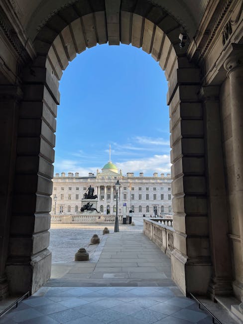 View through a grand stone archway towards the open courtyard of Somerset House, featuring a historic building with a light-colored facade, numerous windows, and a central green-domed roof topped by a